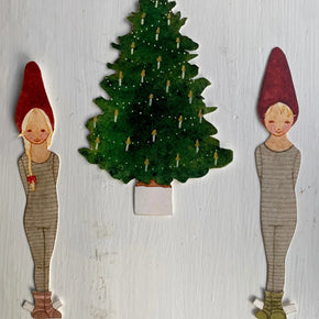 A handmade paper cutout Christmas scene featuring two small gnomes in striped sweaters and red pointed hats flanking a green Christmas tree decorated with candle cutouts, all mounted on a white painted wooden background.