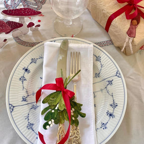 A festive tabletop setting featuring a white plate with blue floral patterns, a white napkin, silver cutlery, a green mistletoe sprig with a red ribbon, a clear glass, and a wrapped gift box on a white tablecloth.