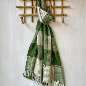 A green and cream plaid woven scarf with fringe tassels, hanging from a bamboo wall hook on a textured off-white wall.