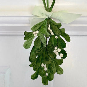 A green mistletoe holiday decoration with white berries and a pale green ribbon bow, hanging on a white paneled door.