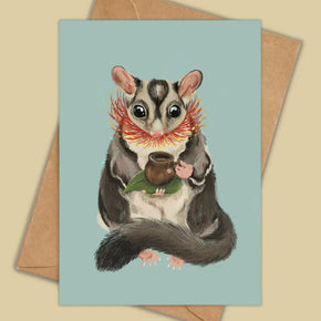 A square greeting card with a light blue background featuring an illustration of a sugar glider holding a small brown cup and green leaf, shown with a kraft paper envelope on a beige background.